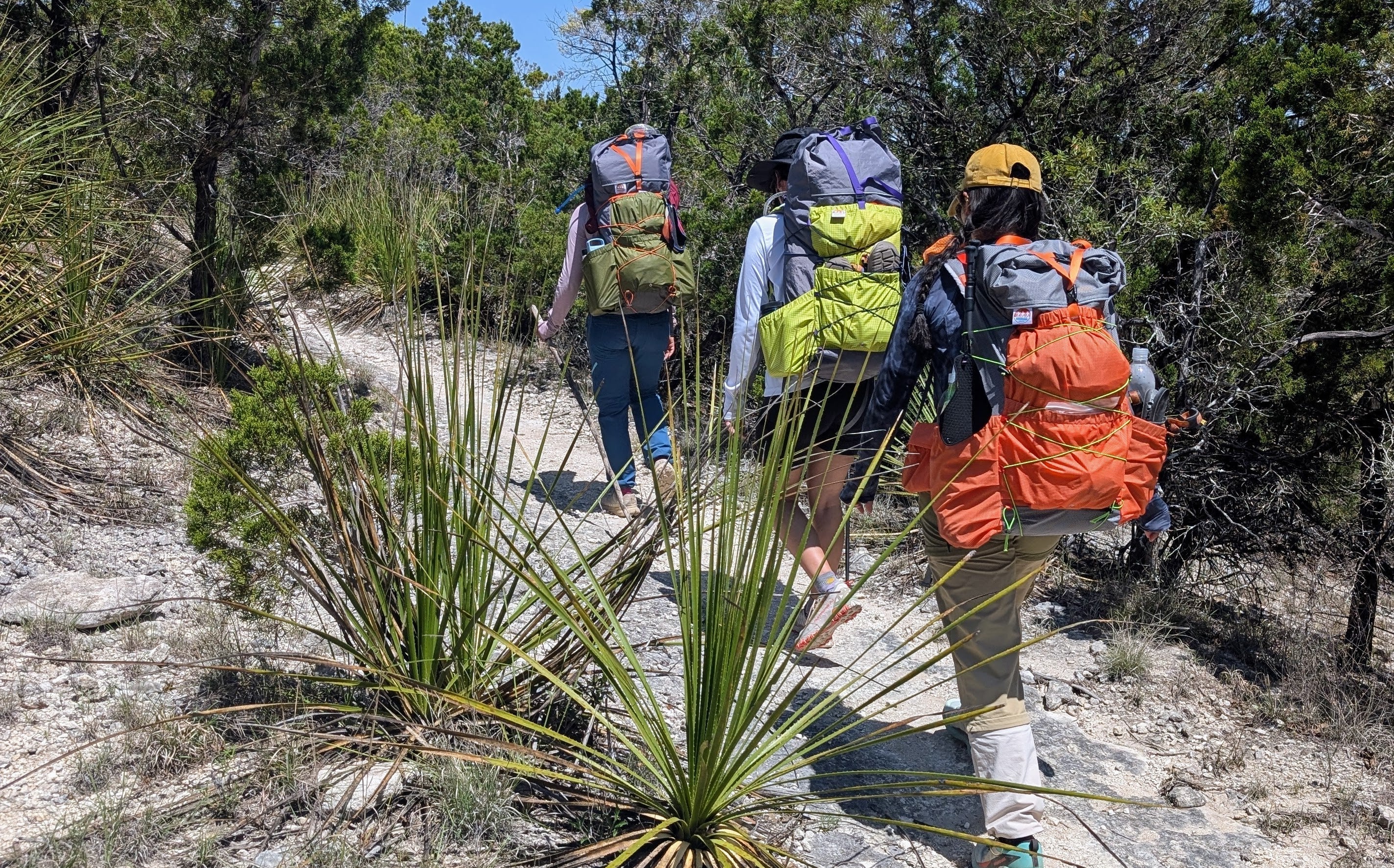3 women backpacking up a hill on rocky terrain wearing Sojourn Trekkr backpacks in the hot summer sun.