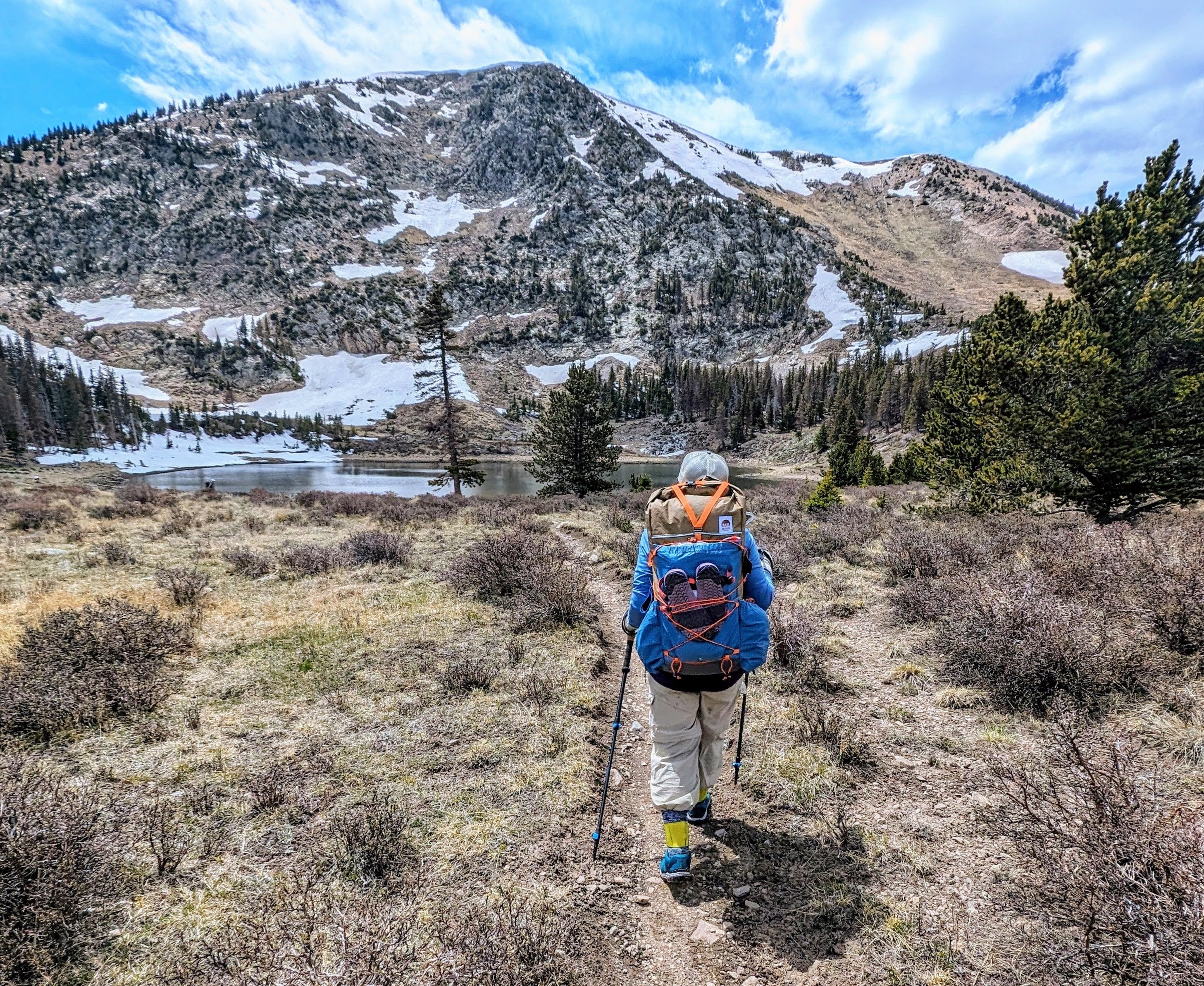 Viewing the back of a solo female backpacker as she hikes towards an alpine lake with a slightly snowy mountain in the background.